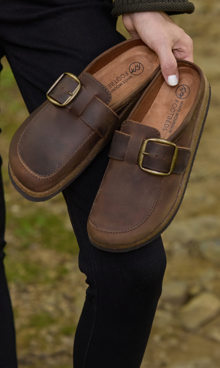 A person holds a pair of brown leather clogs with brass-tone buckles outdoors. The person is wearing black pants, and the background shows a blurred natural stone and grass setting.