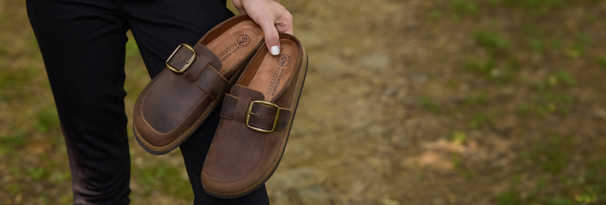 A person outdoors holds a pair of brown leather mule clogs with gold-tone buckles against a blurred dirt and grass background.