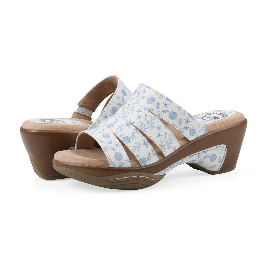 Side view of a pair of open-toe mule sandal in white with a blue seashell pattern, a cushioned insole, and a brown heel 